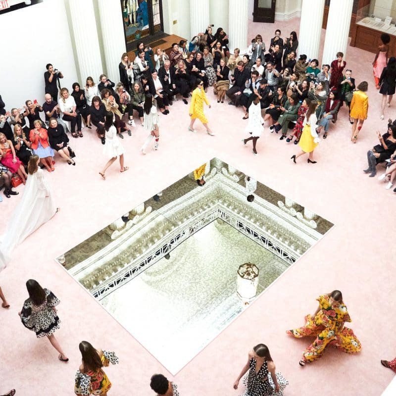 View from above as fashion models circle a large mirror on the floor